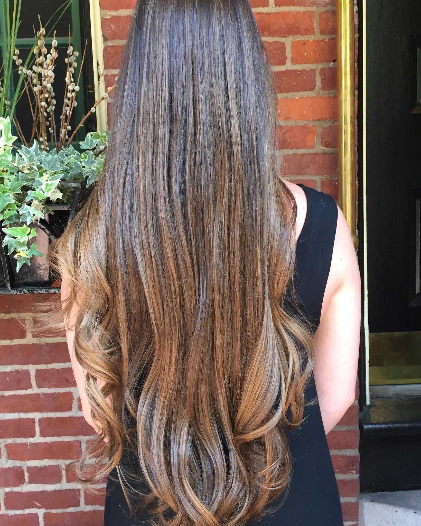 Long, wavy brown hair with highlights, worn by a person in a black top standing against a brick wall.