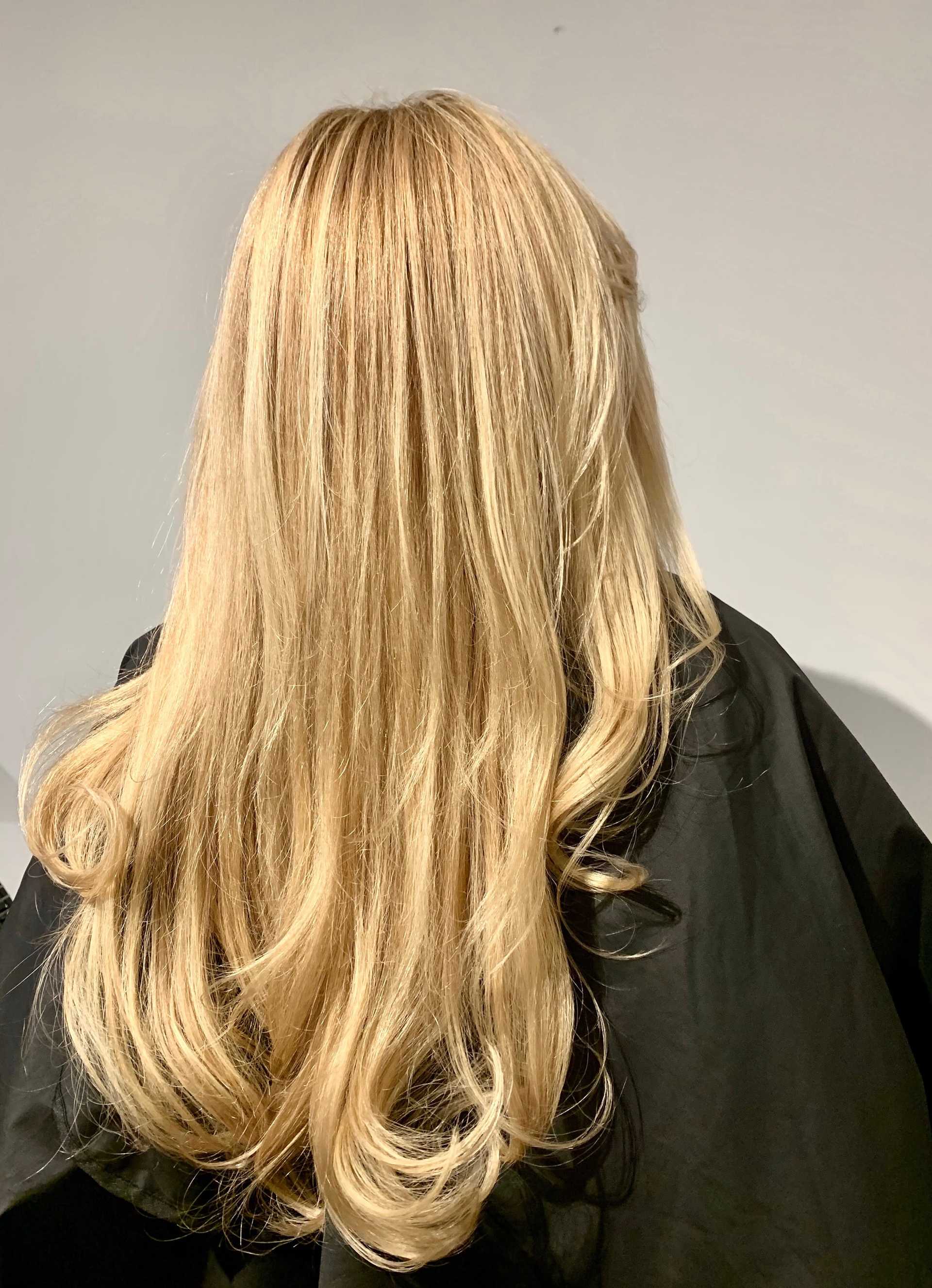 Long blonde hair with soft curls viewed from the back. Black salon cape visible.