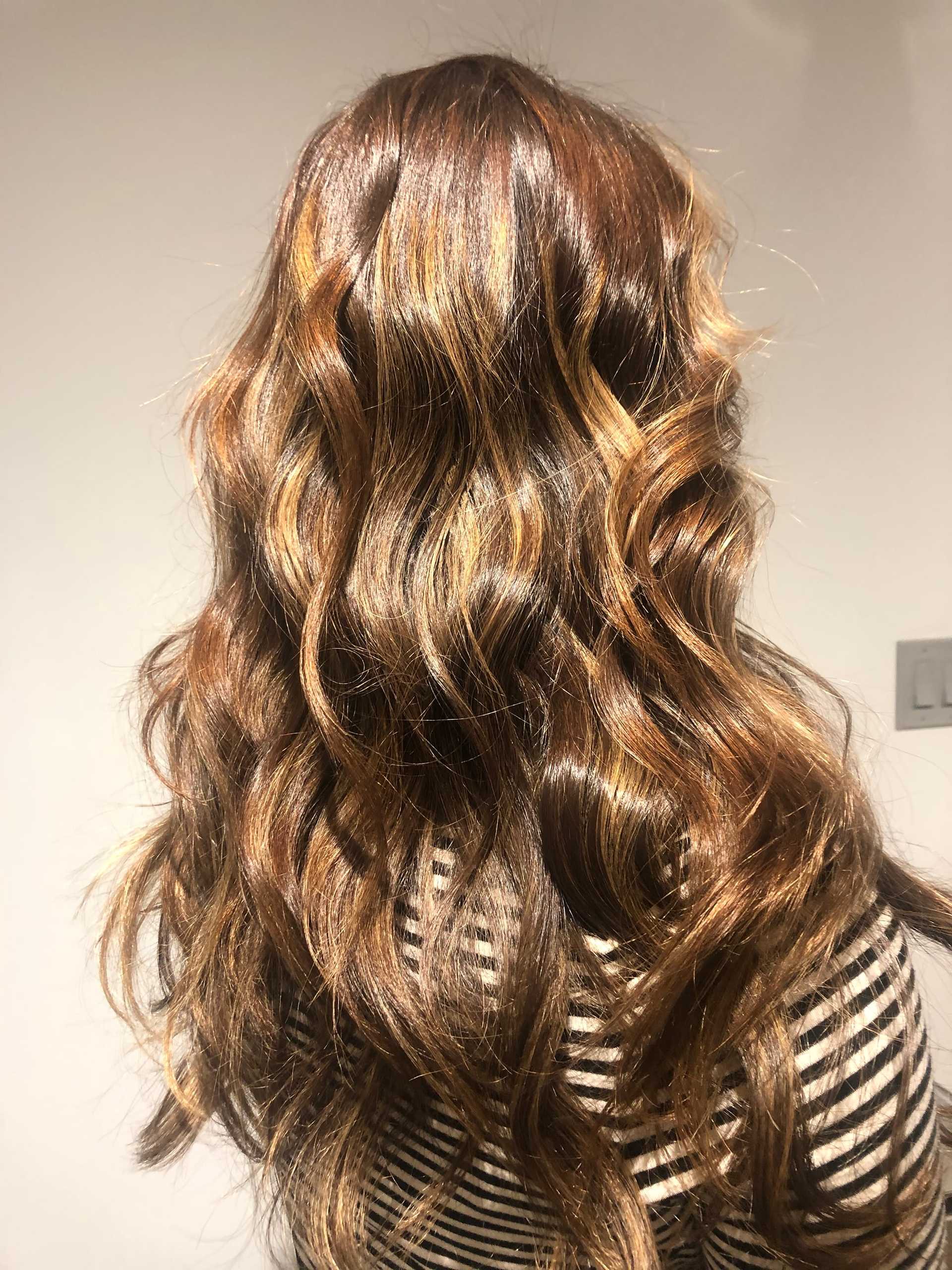Wavy brown hair with blonde highlights cascading over shoulders. Striped top is partially visible.
