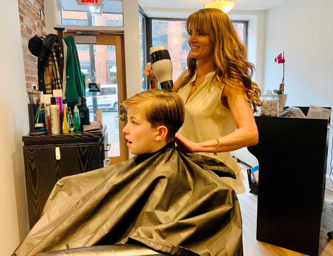 Child getting a haircut at a salon from a smiling hairstylist using a blow dryer.